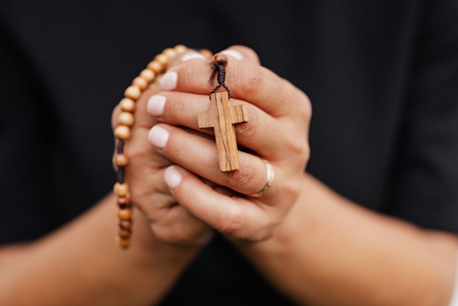 Mulher segurando mãos juntas em oração, com um crucifixo de madeira e um terço, símbolo de fé e espiritualidade.