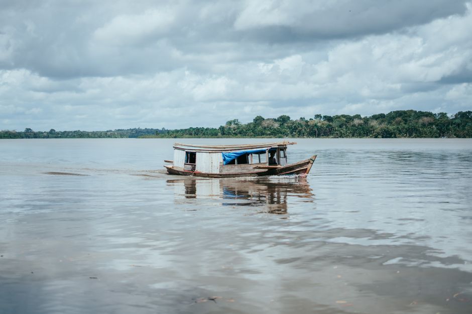 Barco de pesca artesanal navegando em um rio tranquilo, com uma paisagem de floresta ao fundo, simbolizando conexão com a natureza e vida ribeirinha.