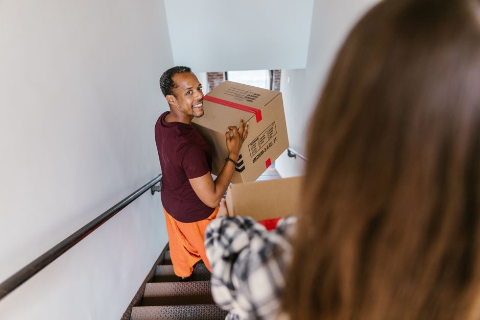 Homem sorridente ajudando a transportar caixas de mudança em escada de prédio para uma breve mudança residencial.