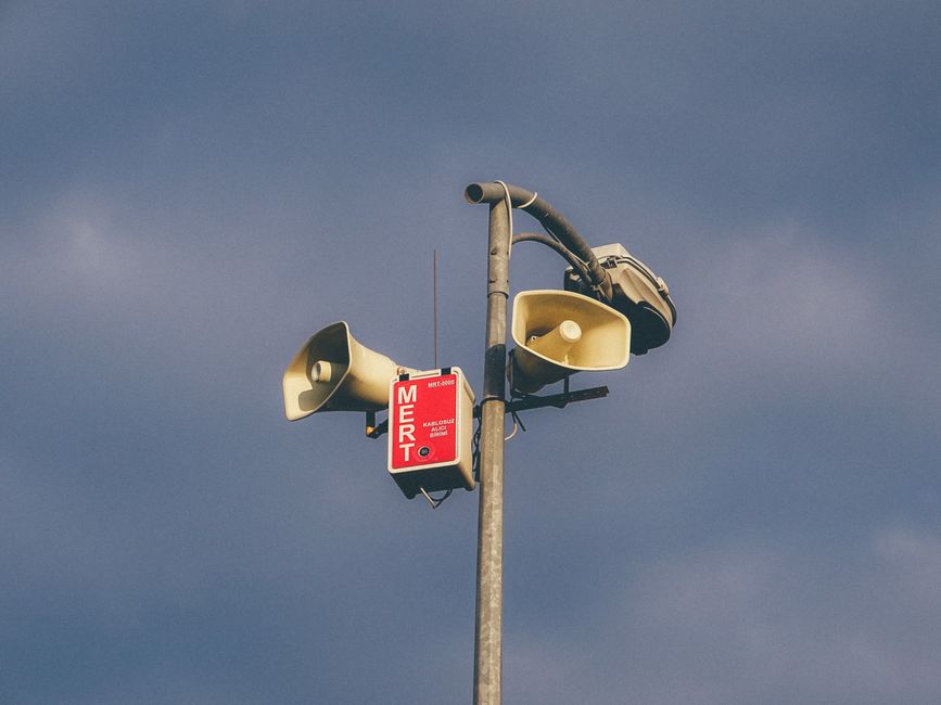 Imagem de um poste de comunicação com alto-falantes e um sinal de advertência, representando alertas e comunicação pública ao ar livre.
