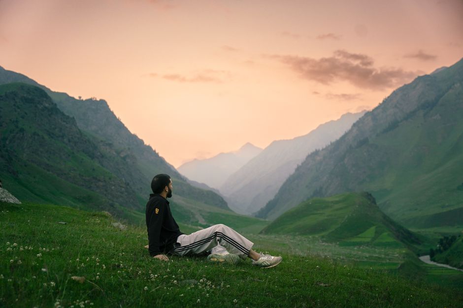 Homem sentado na grama apreciando a paisagem de montanhas ao entardecer, simbolizando tranquilidade e conexão com a natureza.
