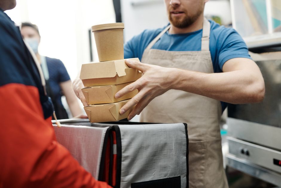 Homem entregando refeições embaladas em papel, representando serviço de entrega de comida ou delivery em ambiente comercial.