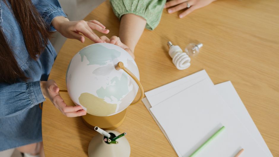 Imagem de uma pessoa ajustando um globo terrestre sobre uma mesa de trabalho, com materiais de escritório ao redor, simbolizando educação, sustentabilidade ou questões ambientais.