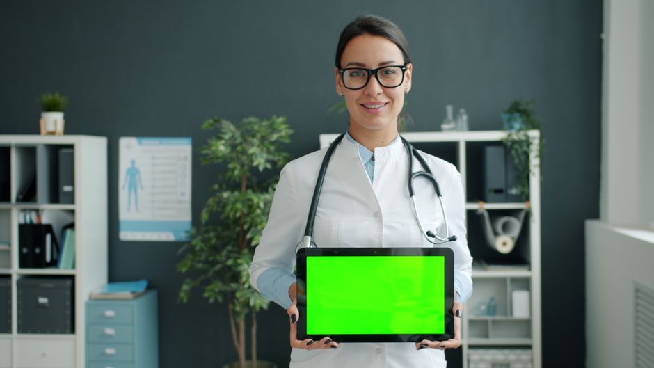 Profissional de saúde mostrando um tablet com tela verde em um ambiente clínico, simbolizando tecnologia na área de saúde.