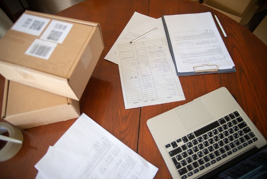 Imagem de uma mesa de trabalho com documentos, fichas de controle, caixas de envio e um laptop, sugerindo organização e processos administrativos ou de comércio eletrônico.