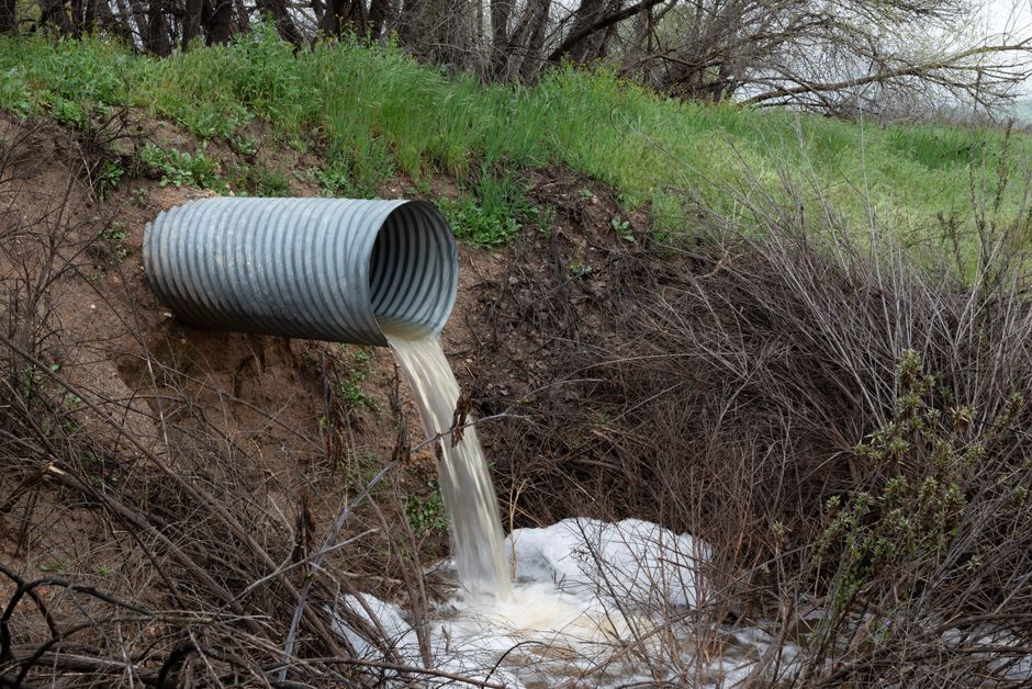 Imagem de um tubo de metal despejando água barrenta em um riacho, simbolizando poluição ambiental e impacto na natureza.