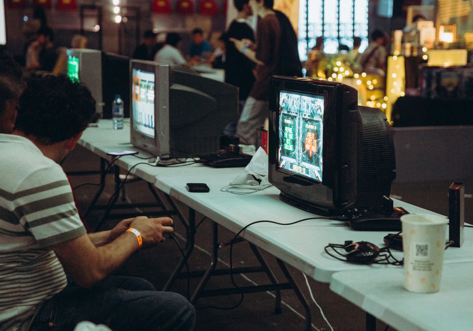 Homem jogando videogame em um evento de competição ou lan party, com várias telas e outros participantes ao fundo.