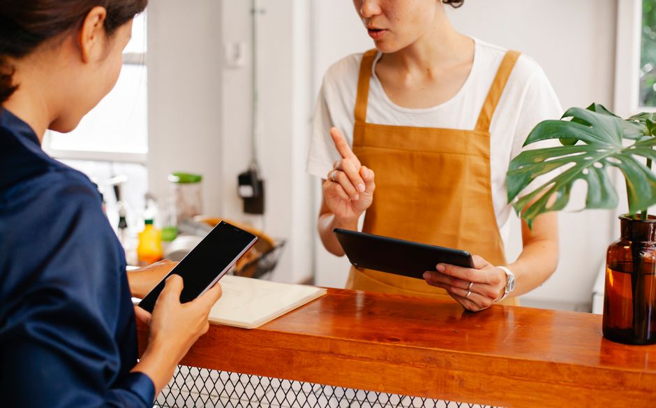 Uma atendente de restaurante ou cafeteria interagindo com uma cliente na bancada, utilizando tablet e smartphone para gerenciar pedidos ou pagamentos.