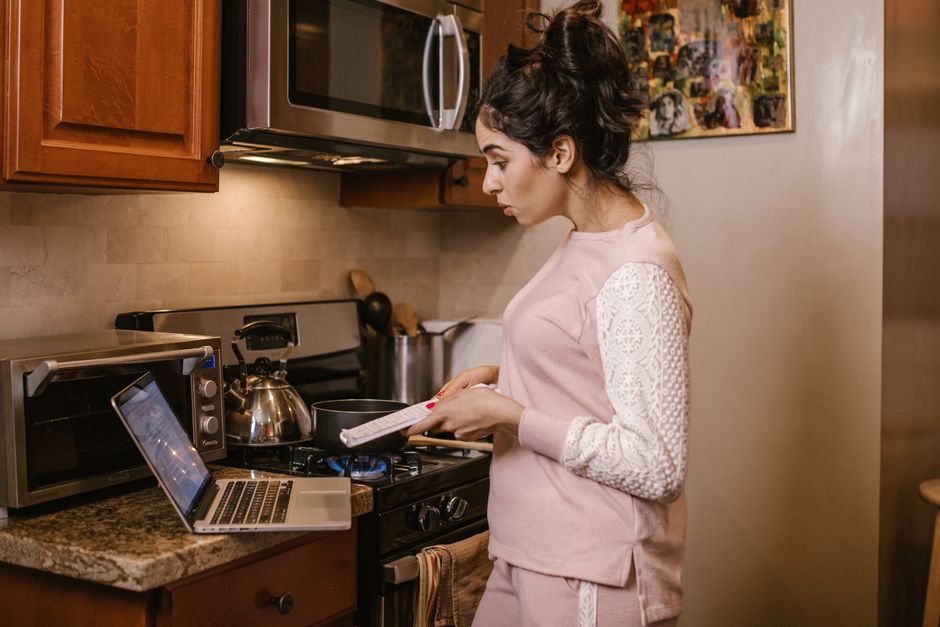 Jovem mulher usando notebook e telefone enquanto cozinha na cozinha de sua casa, combinando tecnologia e tarefas domésticas.