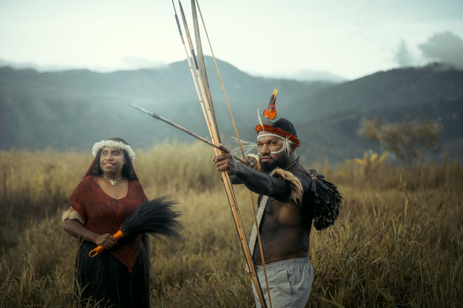 Homens indígenas tradicionais em vestimenta ceremonial, com cenário natural de montanhas ao fundo.