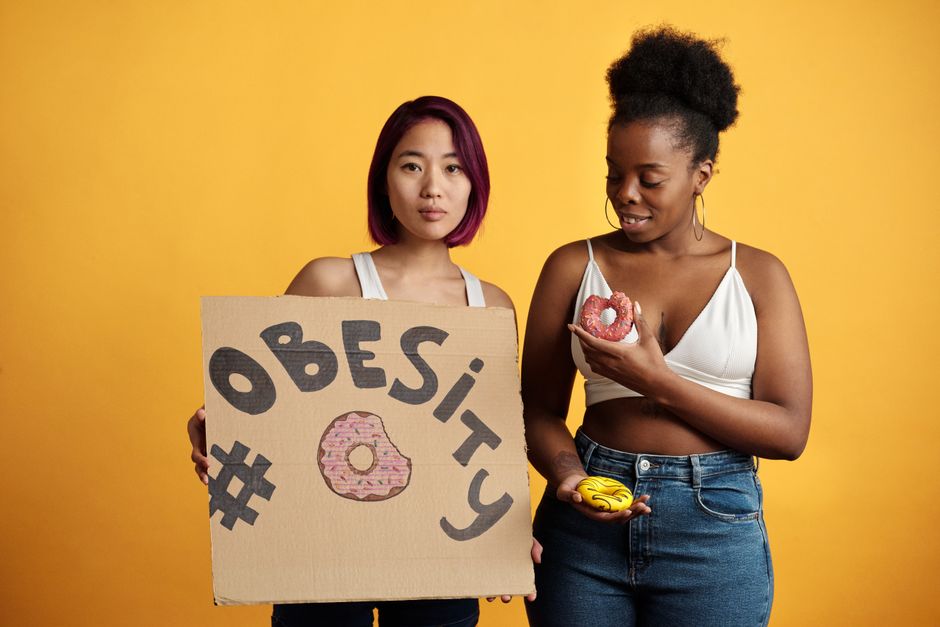 Duas mulheres segurando sinal e alimentos coloridos, abordando o tema da obesidade e suas questões relacionadas.