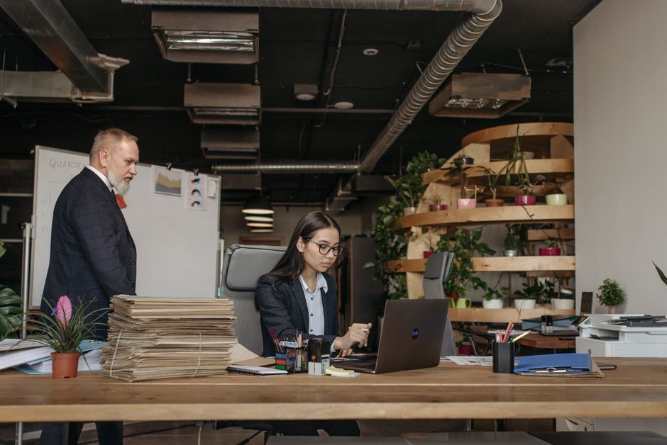Imagem de um escritório moderno com uma mulher usando laptop e um homem de terno observando ao fundo, destacando o ambiente de trabalho e gestão empresarial.