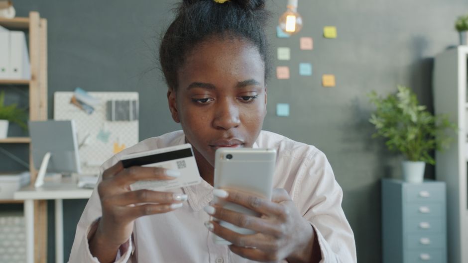 Mulher jovem realizando uma compra online usando cartão de crédito e celular em ambiente de escritório moderno.
