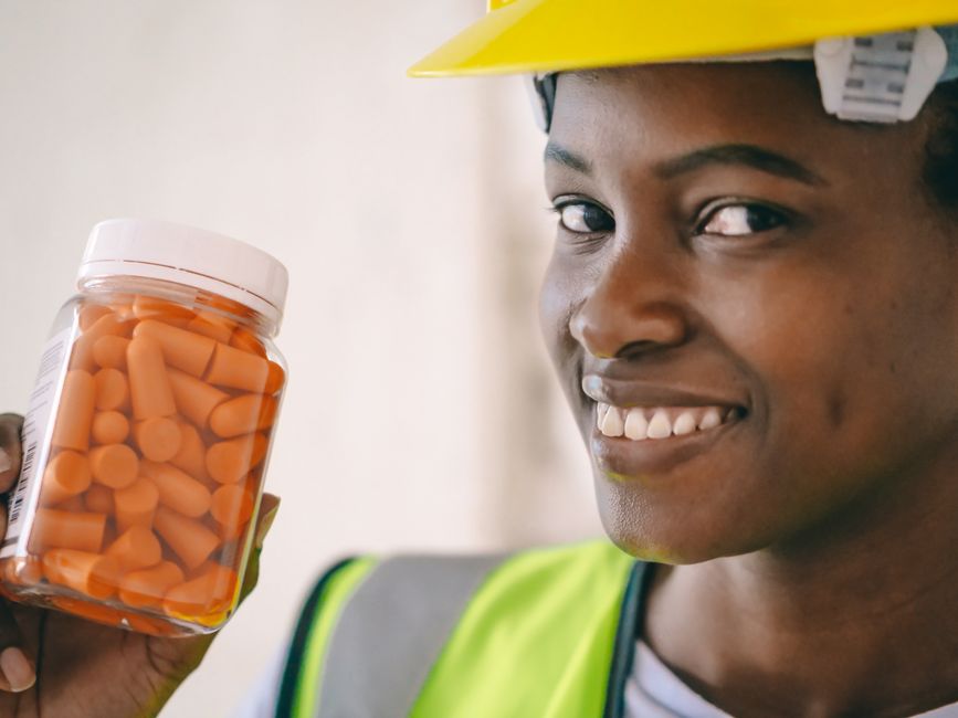 Mulher sorridente usando capacete de segurança e vestimenta de trabalho, segurando uma jarra com comprimidos, símbolo de saúde e cuidados medicamentosos.