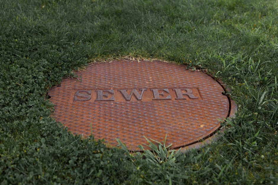 Imagem de uma tampa de bueiro de ferro com a palavra 'SEWER' instalada no gramado, simbolizando infraestrutura urbana de saneamento básico.