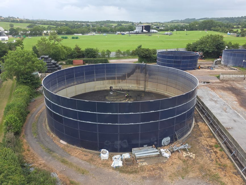 Imagem aérea de uma instalação industrial com tanques de armazenamento em meio a uma paisagem rural com áreas verdes e um palco ao fundo.