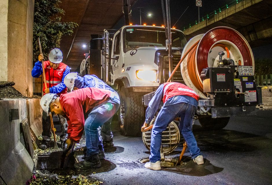 Equipe de trabalhadores realizando manutenção na infraestrutura urbana sob uma ponte à noite, com uma viatura de serviços ao fundo.