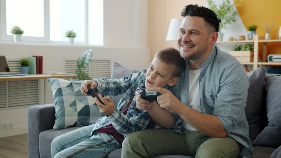 Pai e filho se divertem jogando video game juntos no sofá de um ambiente aconchegante e bem iluminado.