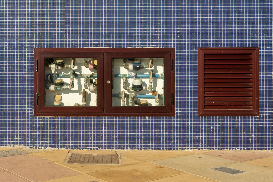 Vista de uma parede de prédio com duas janelas de vidro que mostram tubulações e componentes hidráulicos internos, refletindo uma estrutura de manutenção predial.