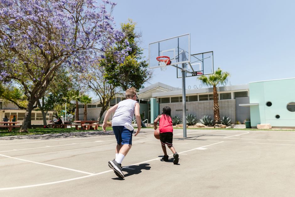 Crianças brincando de basquete em quadra ao ar livre em um dia ensolarado, com árvores floridas ao redor.