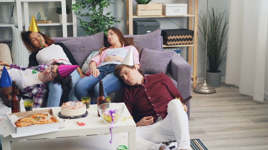 Grupo de amigos dormindo e descansando após uma festa, com decorações de chapéus e festa na sala de estar.