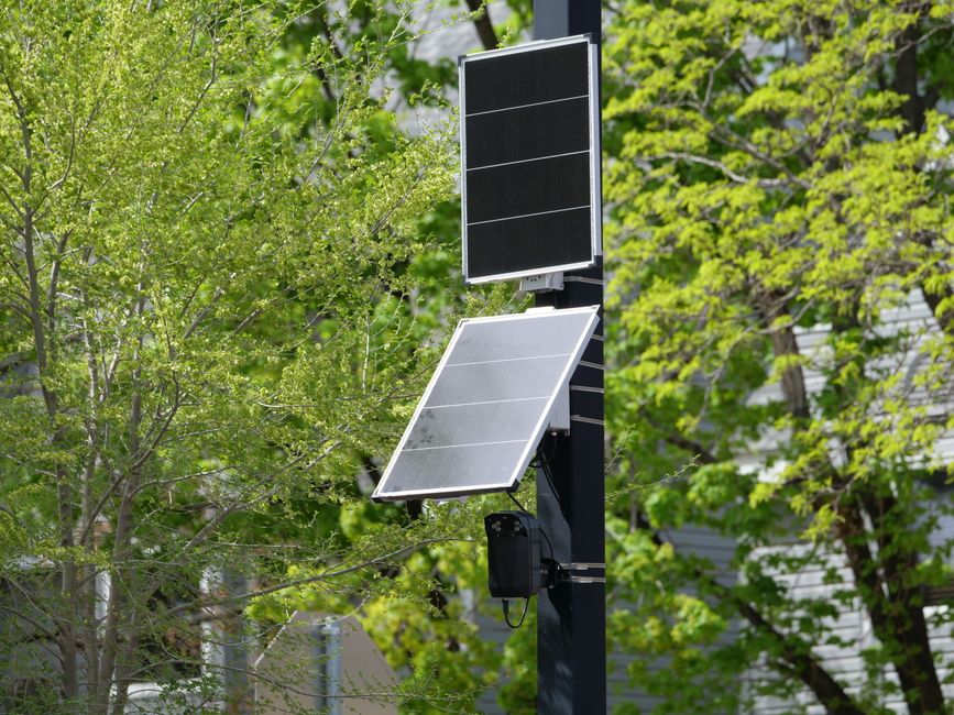 Poste de energia solar com painéis fotovoltaicos instalado em área urbana com árvores verdes ao fundo, representando sustentabilidade e energia renovável.
