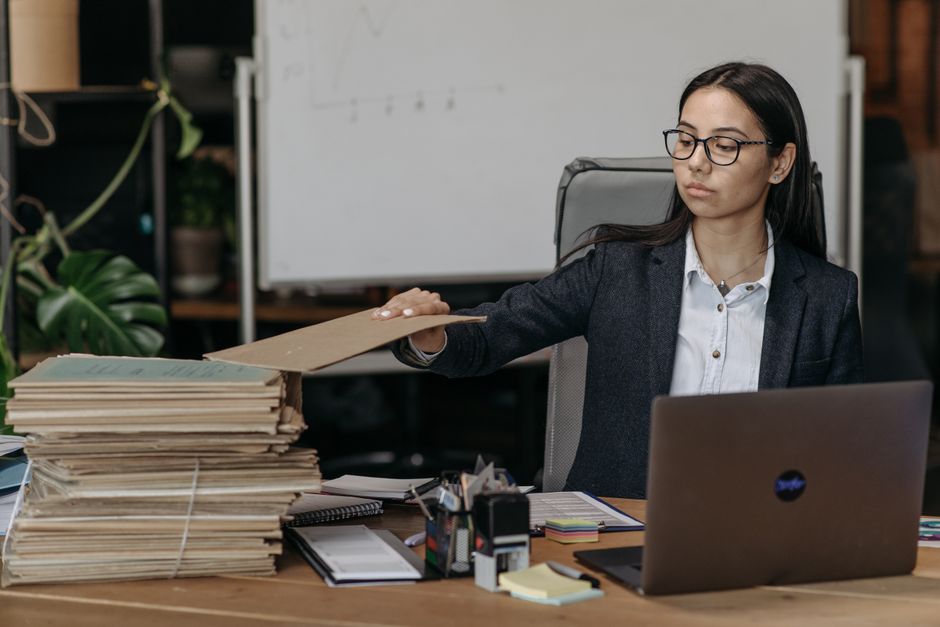Imagem de uma mulher de óculos e blazer organizando uma pilha de documentos em um ambiente de escritório, indicando rotina de trabalho burocrática ou de gestão de arquivos.