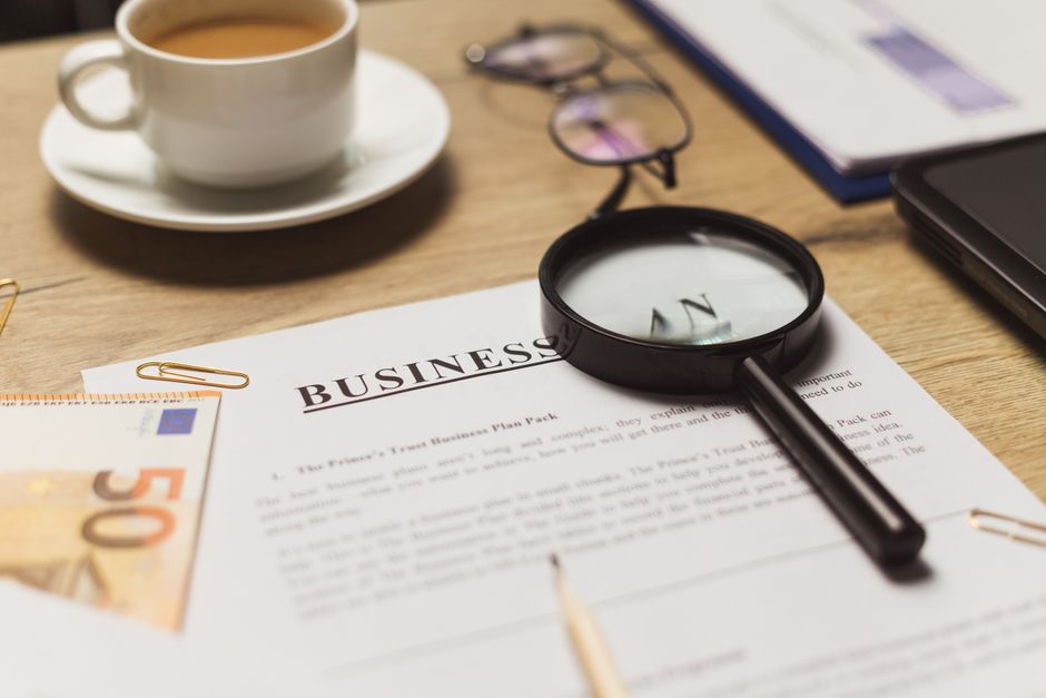 Imagem de uma mesa de trabalho com documentos de negócios, uma xícara de café, óculos, uma lupa e elementos financeiros, refletindo planejamento e análise empresarial.
