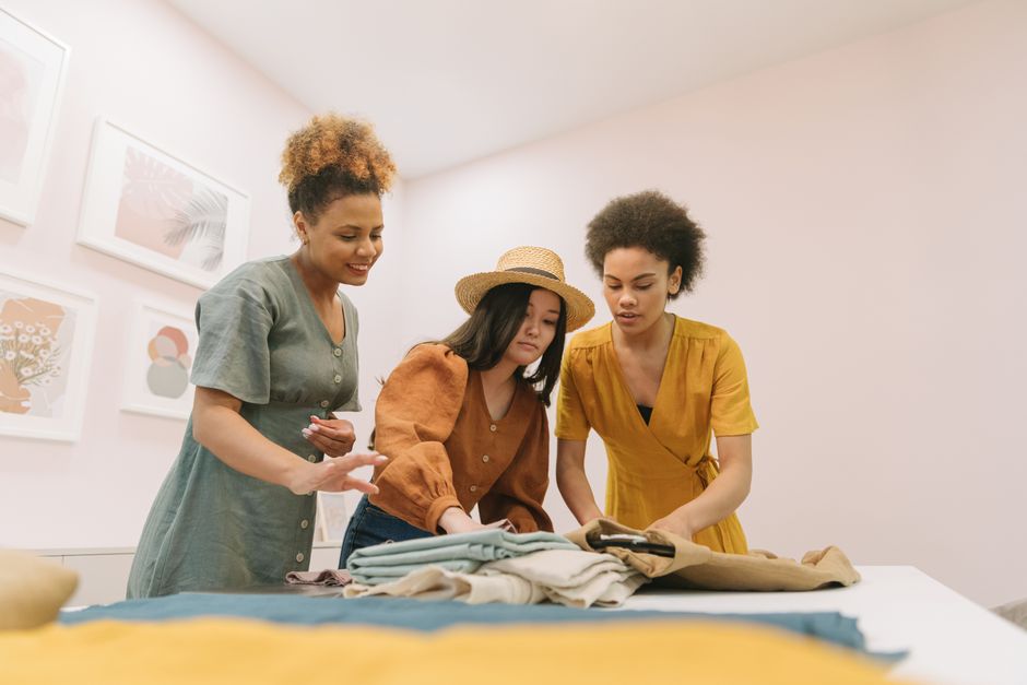 Três mulheres discutindo roupas e estilo em um ambiente de loja ou atelier de moda, focando na criatividade e tendências na moda feminina.
