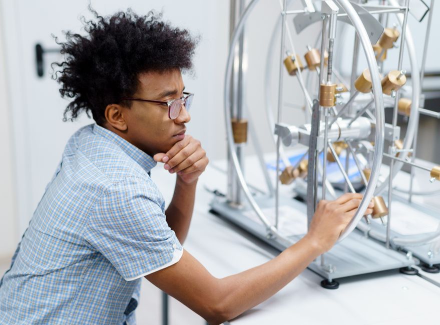 Estudante concentrado, observando e ajustando um dispositivo de pesquisa ou experimento científico em laboratório.