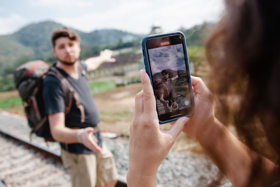 Pessoa gravando vídeo de uma outra pessoa ao ar livre, próximo a trilhos de trem, com cenário natural de montanhas ao fundo.