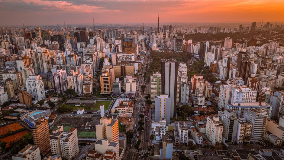 Imagem de uma vista panorâmica de uma cidade grande ao pôr do sol, destacando o centro urbano com edifícios altos e ruas movimentadas.