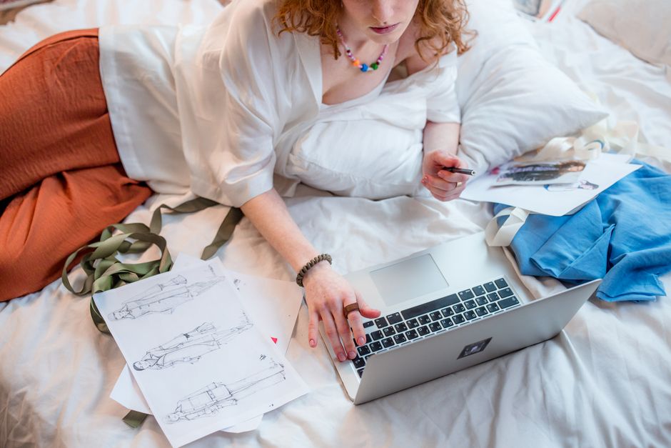Mulher deitada na cama trabalhando com documentos de moda e usando laptop, em um ambiente casual e criativo.