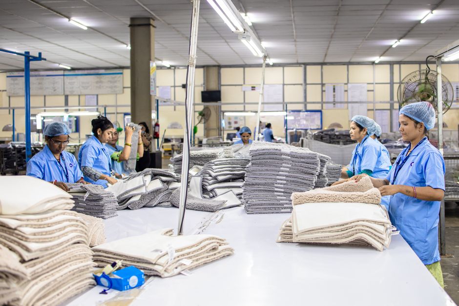 Imagem de uma fábrica têxtil com operárias organizando e conferindo tecidos e roupas em um ambiente industrial bem iluminado.