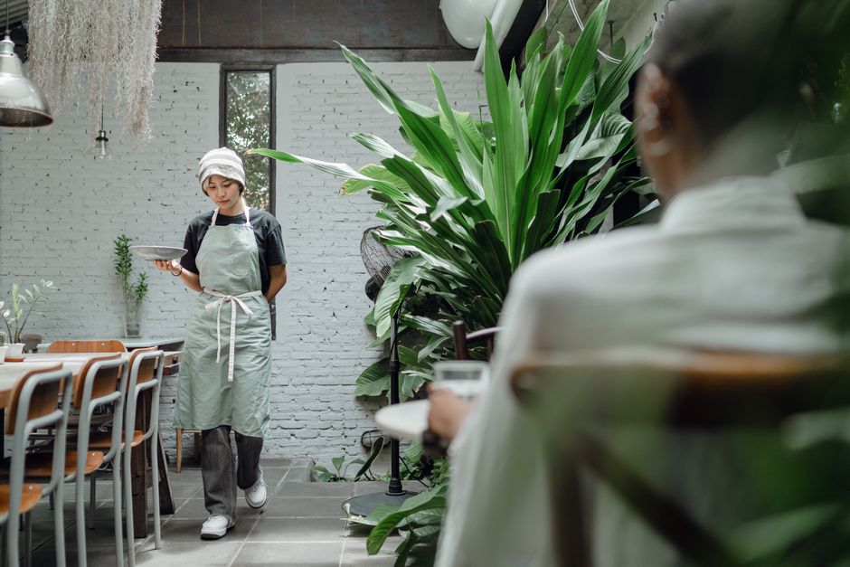 Imagem de um ambiente de restaurante com uma garçonete verificando um dispositivo enquanto uma cliente observa, com elementos decorativos de plantas naturais ao fundo.