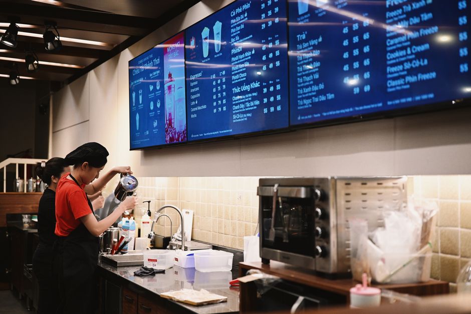Funcionárias preparando bebidas em uma cafeteria moderna, com painel digital de cardápio ao fundo.