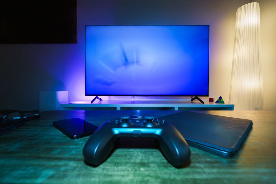 Imagem de uma mesa de entretenimento com um controle de videogame, uma câmera e um notebook, com uma televisão moderna ao fundo, ideal para artigos sobre jogos e tecnologia.