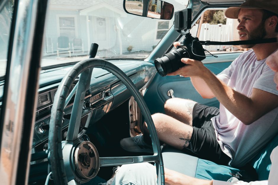 Homem no banco do passageiro utilizando câmera profissional enquanto outro está ao volante de um veículo em ambiente residencial.