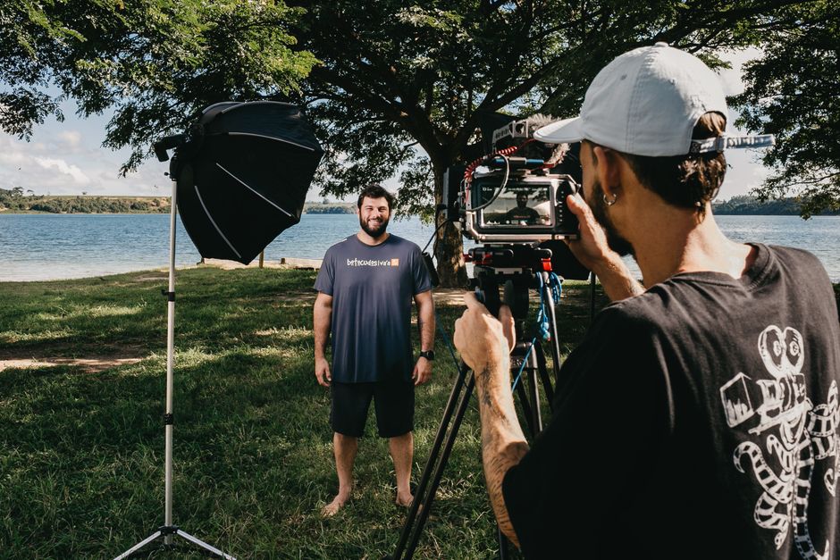 Pessoa gravando entrevista ou vídeo ao ar livre com equipamento de filmagem próximo a rio ou lago, em ambiente natural com árvores e céu parcialmente nublado.