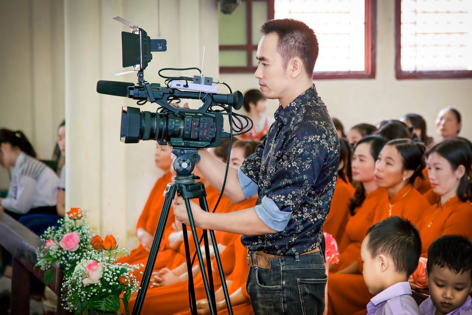 Homem gravando uma cerimônia ou evento cultural com câmera profissional, em ambiente de sala de aula ou auditório cheio de pessoas vestidas de forma tradicional ou formal.