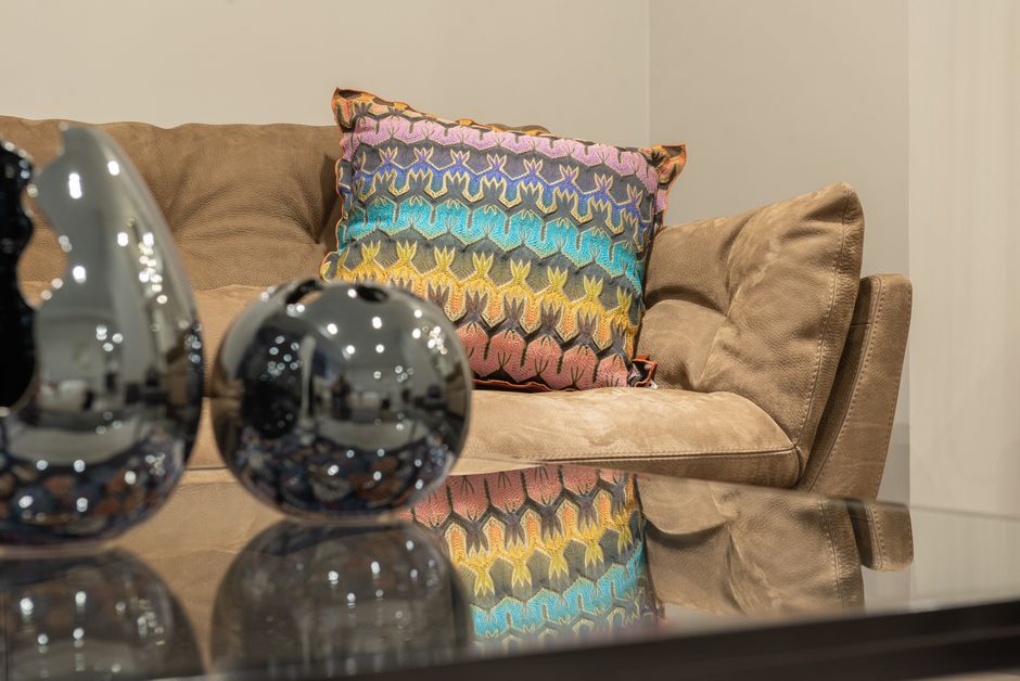 Imagem de uma sala de estar aconchegante com um sofá confortável, uma almofada decorativa colorida e objetos decorativos de metal, transmitindo um ambiente receptivo e elegante.