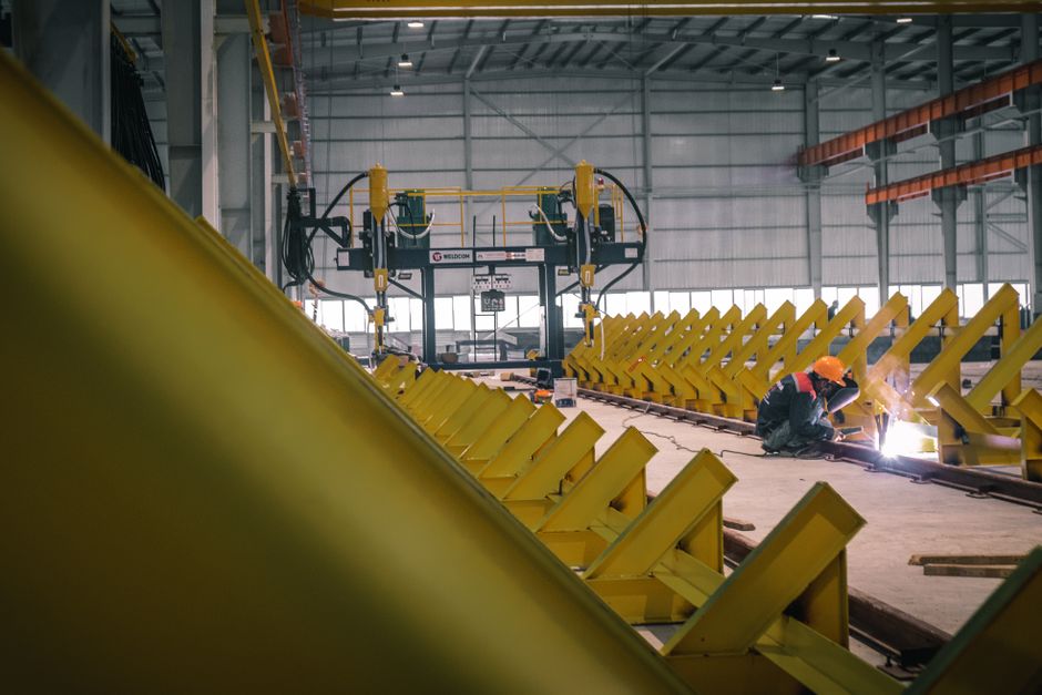 Imagem de uma fábrica industrial com trabalhador realizando soldagem em estrutura metálica, destacando processos de fabricação e montagem de equipamentos pesados.