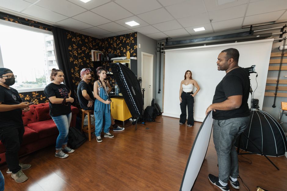 uma sessão de fotos em estúdio com modelo posando diante de um fundo branco, enquanto o fotógrafo orienta e prepara o equipamento para a cobertura fotográfica