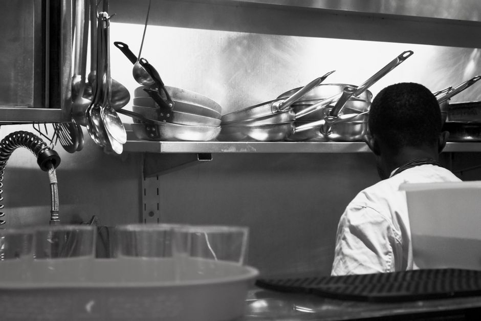Imagem de um profissional de cozinha visto de trás, organizando utensílios em uma cozinha industrial.