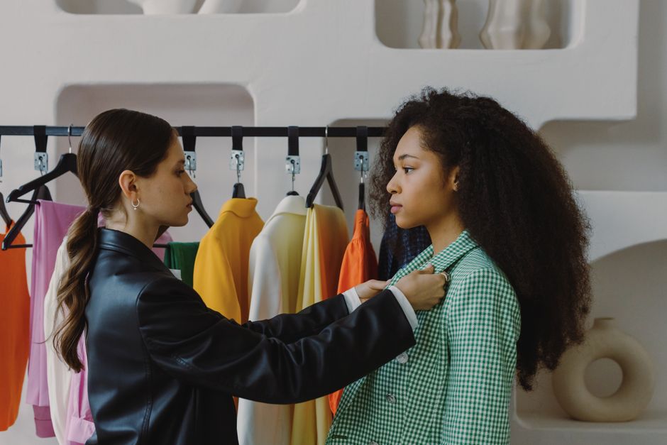 Duas mulheres em uma loja de roupas, uma delas ajudando a ajustar a blazer na outra, representando atendimento ao cliente e moda feminina.