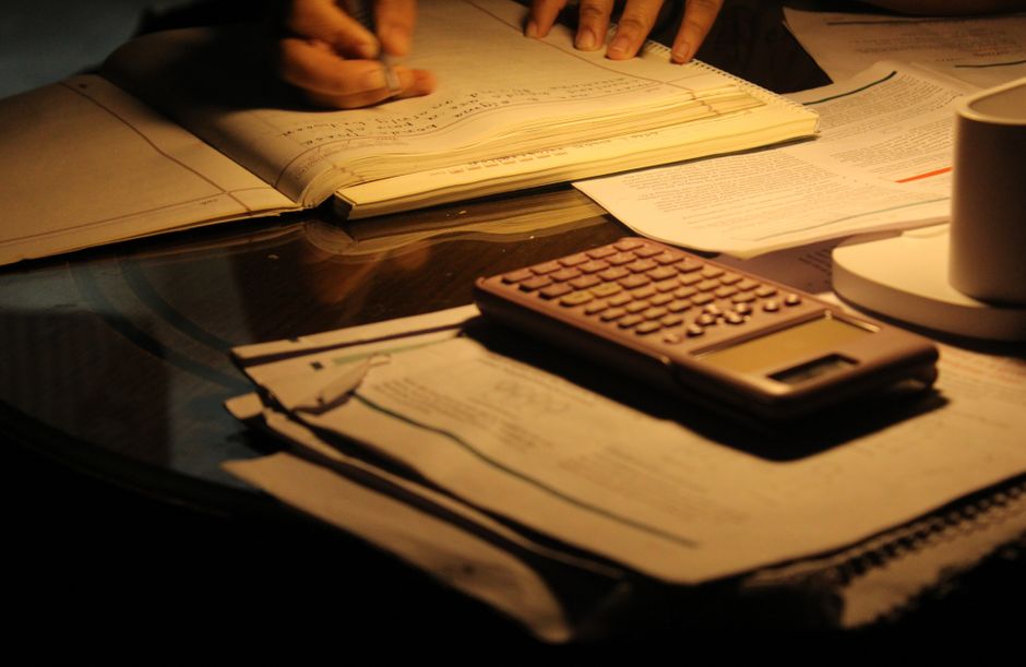 Imagem de uma mesa de trabalho com documentos, calculadora e uma pessoa revisando papéis, em um ambiente de estudo ou escritório.