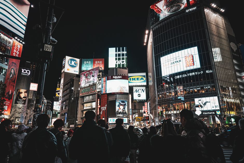 Imagem de uma movimentada rua urbana à noite, com grandes edifícios iluminados e painéis de LED, refletindo o dinamismo e a modernidade da cidade.