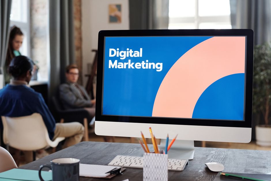 Imagem de um ambiente de trabalho com foco em uma tela de computador exibindo o tema marketing digital, ao fundo, pessoas participando de uma reunião ou palestra.