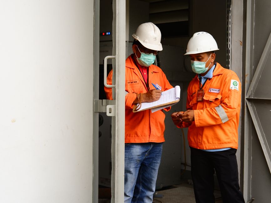 Profissionais de engenharia em fiscalização de obra, utilizando EPIs e realizando anotações em ambiente industrial.