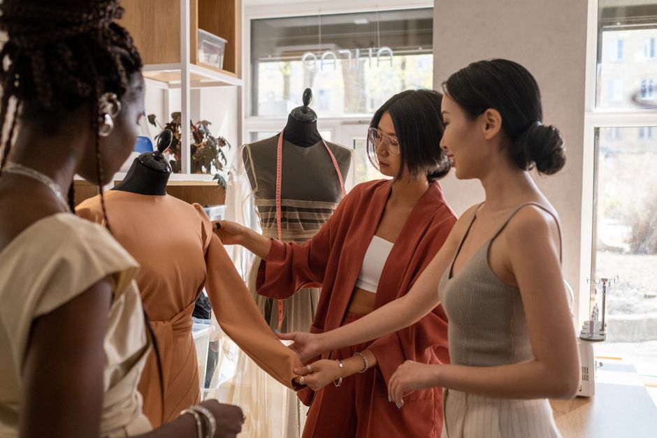 Três mulheres trabalhando juntas na criação e ajuste de roupas em um ateliê de moda, discutindo detalhes de um vestido de cós largo e mangas bufantes.